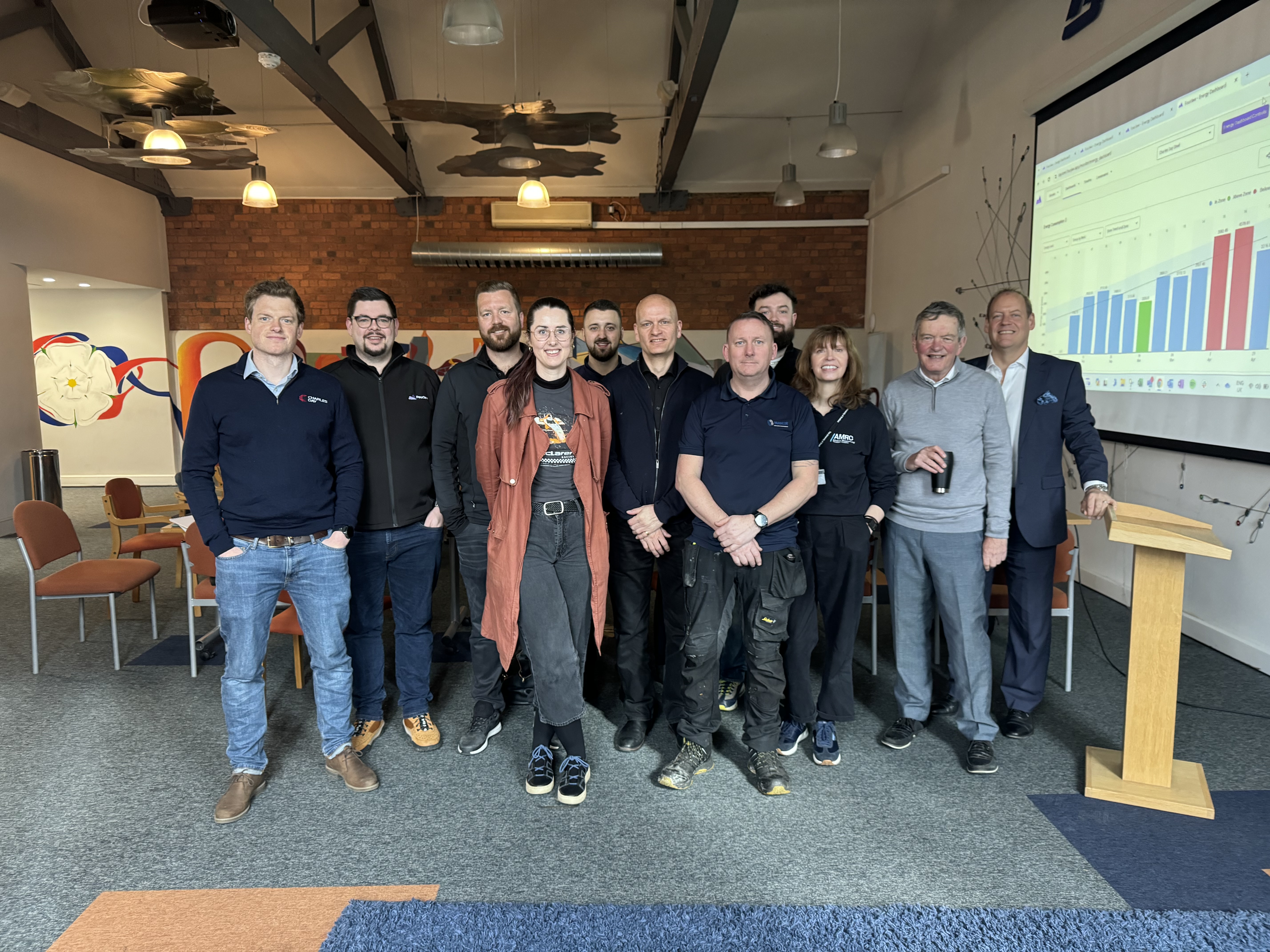 A group of people standing together in a modern meeting room with exposed beams and brick walls. There are orange chairs arranged around the room and a wooden podium on the right. A large screen displays a colourful bar chart with multiple data points. The setting appears to be a business or training event.