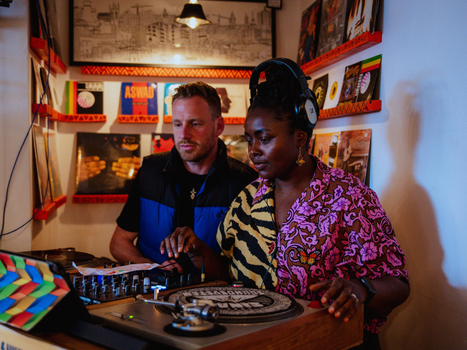Two people standing at a DJ booth inside a room decorated with vinyl records on shelves. One person is wearing headphones and a vibrant pink patterned shirt with a zebra-print scarf, while the other is adjusting controls on the mixer. A turntable with a colorful slipmat is in the foreground, and framed artwork hangs on the wall above.