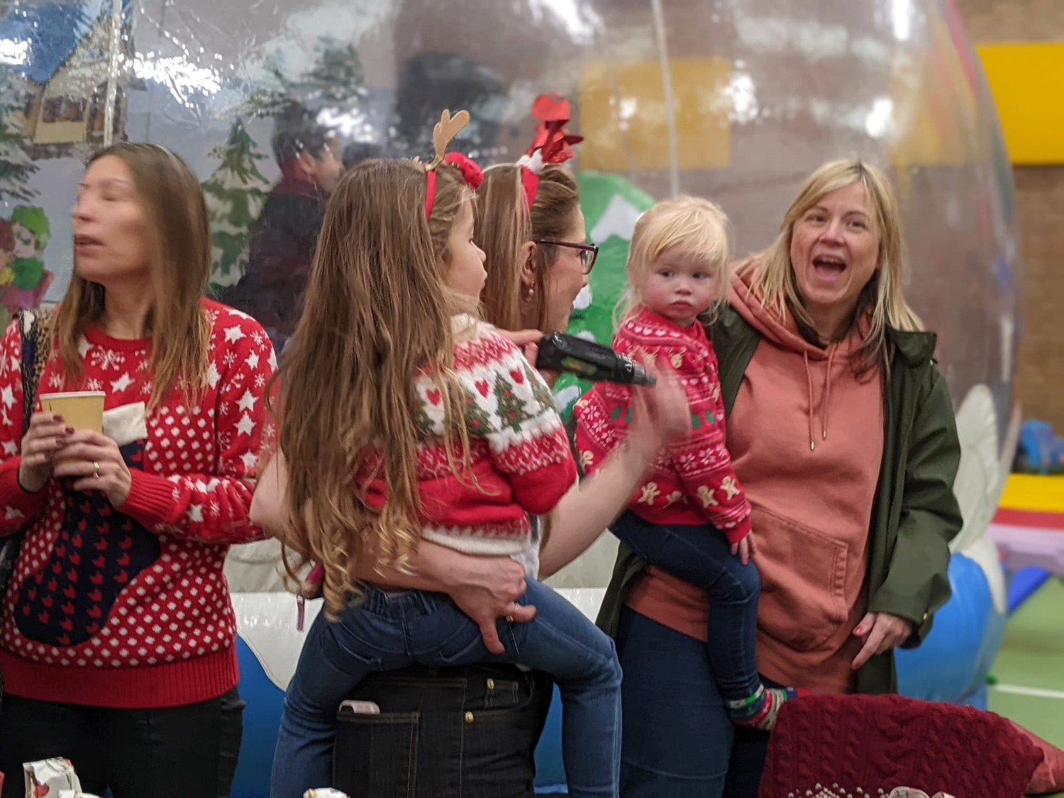 Group of people wearing festive Christmas sweaters, some with reindeer antlers headbands, standing indoors near a large decorative snow globe. Two children are being held by adults, and one person is holding a drink cup. The background includes colourful inflatable decorations and a winter-themed backdrop inside the snow globe.