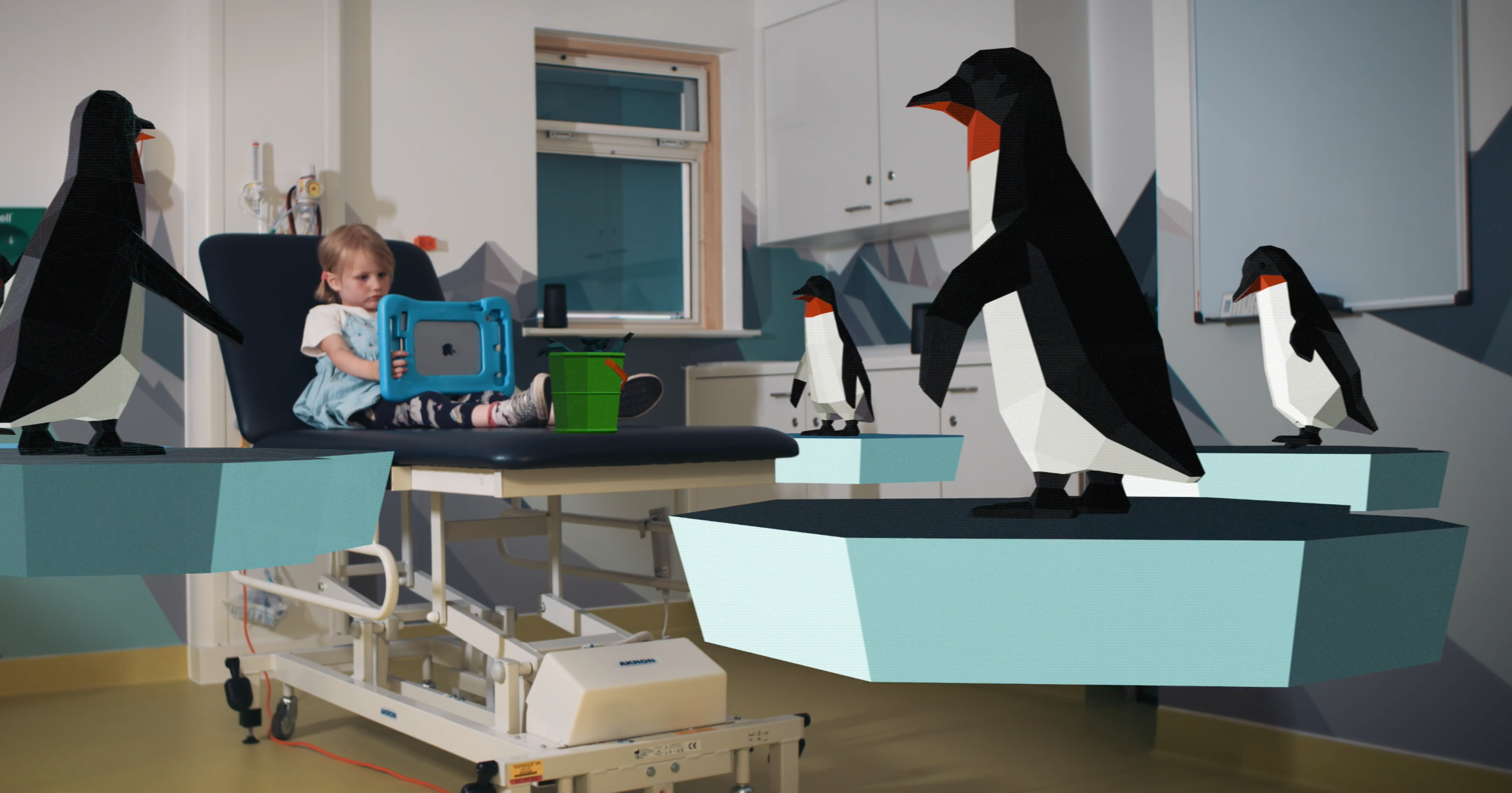 A child is lying in a bed in a hospital ward. There are 3D models of penguins around the room. The child is entertaining herself with a tablet type device.