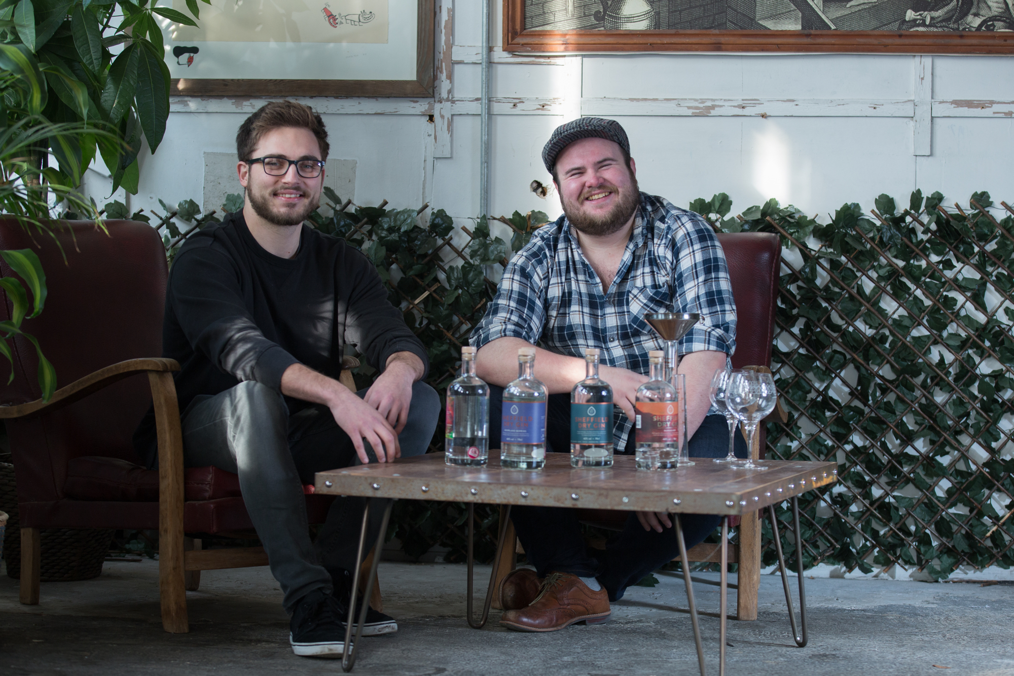 Two people seated on armchairs at a rustic wooden table displaying several bottles of spirits, a cocktail shaker, and empty glasses. The setting includes a leafy backdrop, framed artwork on the wall, and natural light creating a casual, relaxed atmosphere.