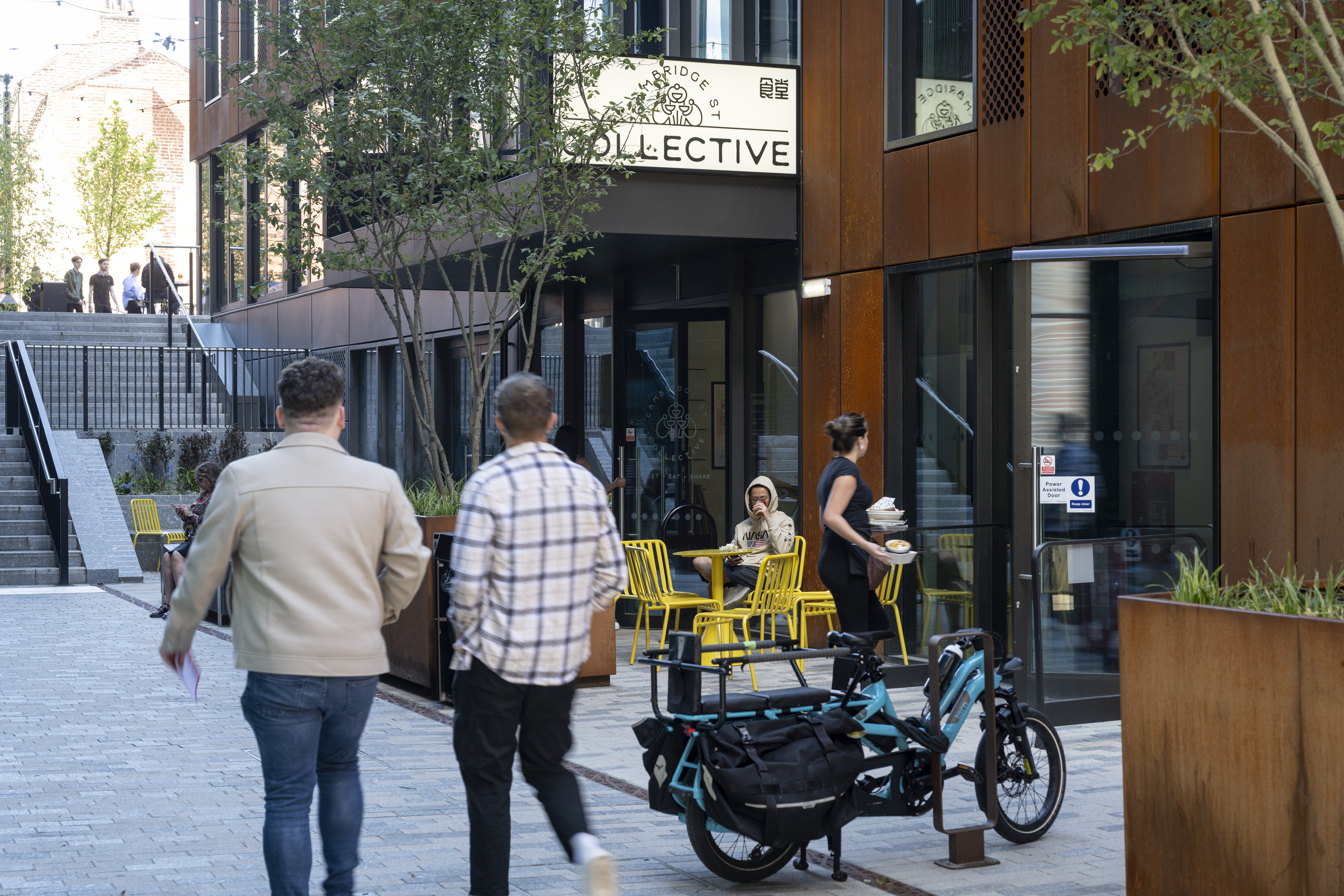Two people walking along a paved pedestrian street lined with modern buildings featuring large windows and rust-colored panels. In the foreground, a cargo-style electric bike is parked near a planter. To the right, a café with bright yellow chairs and tables is visible, with one person seated and another carrying drinks. A sign above the café entrance reads “COLLECTIVE.” Trees and steps leading to an upper level appear in the background.
