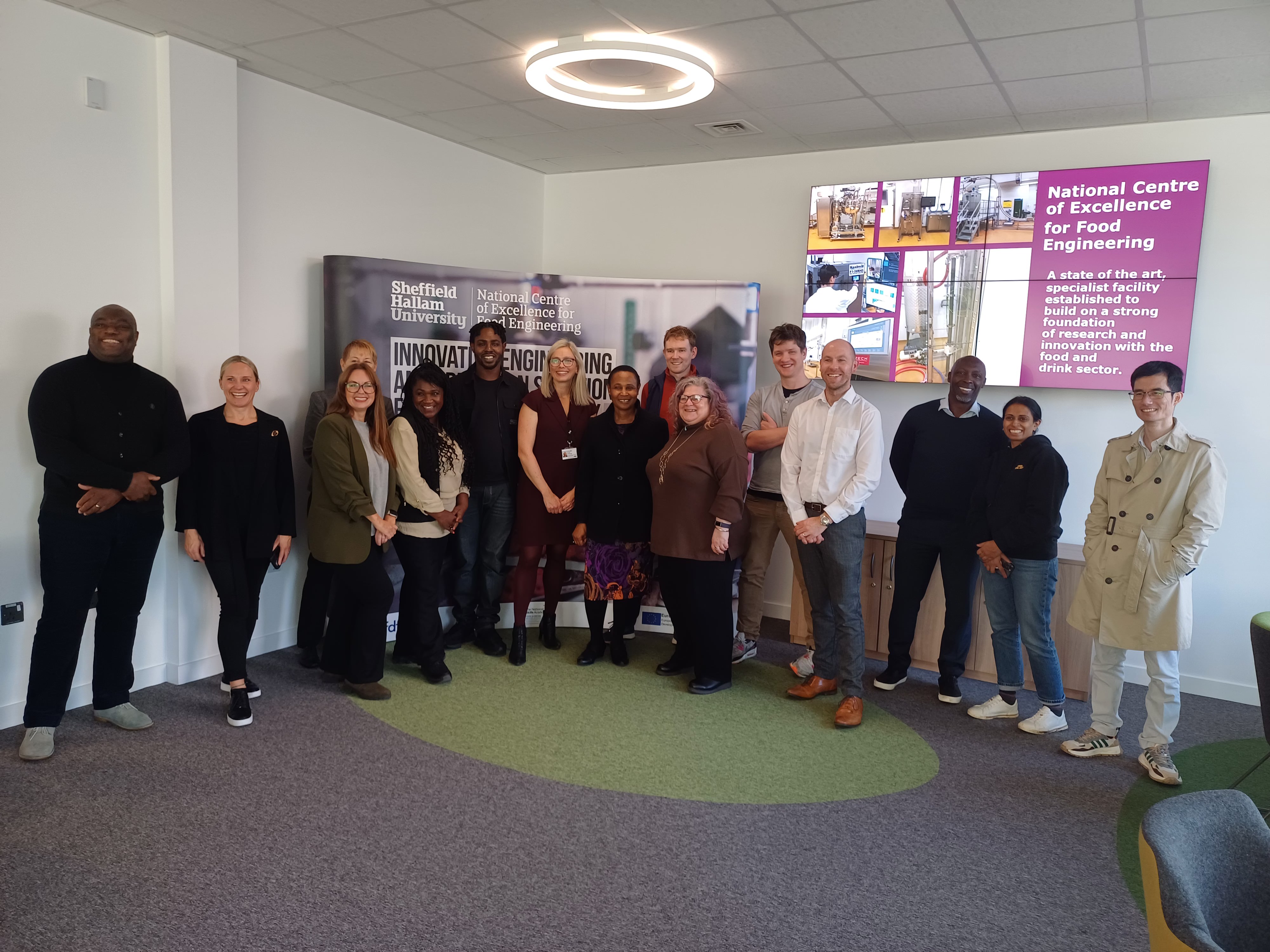 A group of twelve people standing in a semi-circle inside a modern meeting room with grey carpet and green accents. Behind them is a large banner displaying “Sheffield Hallam University” and “National Centre of Excellence for Food Engineering.” On the right wall, there is a digital screen showing images and text about the centre. The room has white walls, a ceiling light, and a few chairs visible on the side.