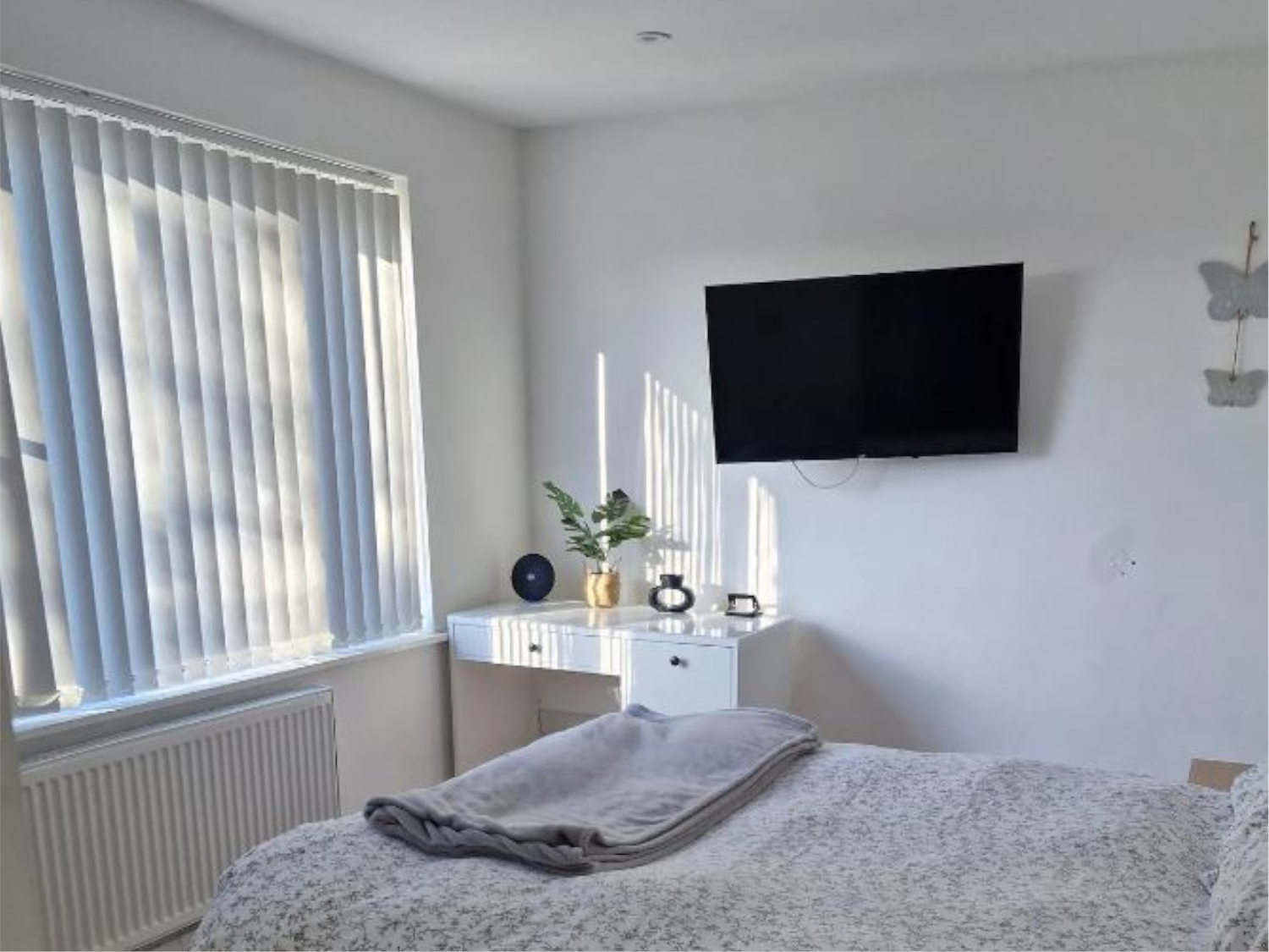 A modern bedroom featuring light linen, a wall-mounted flat-screen TV, a desk with a window.