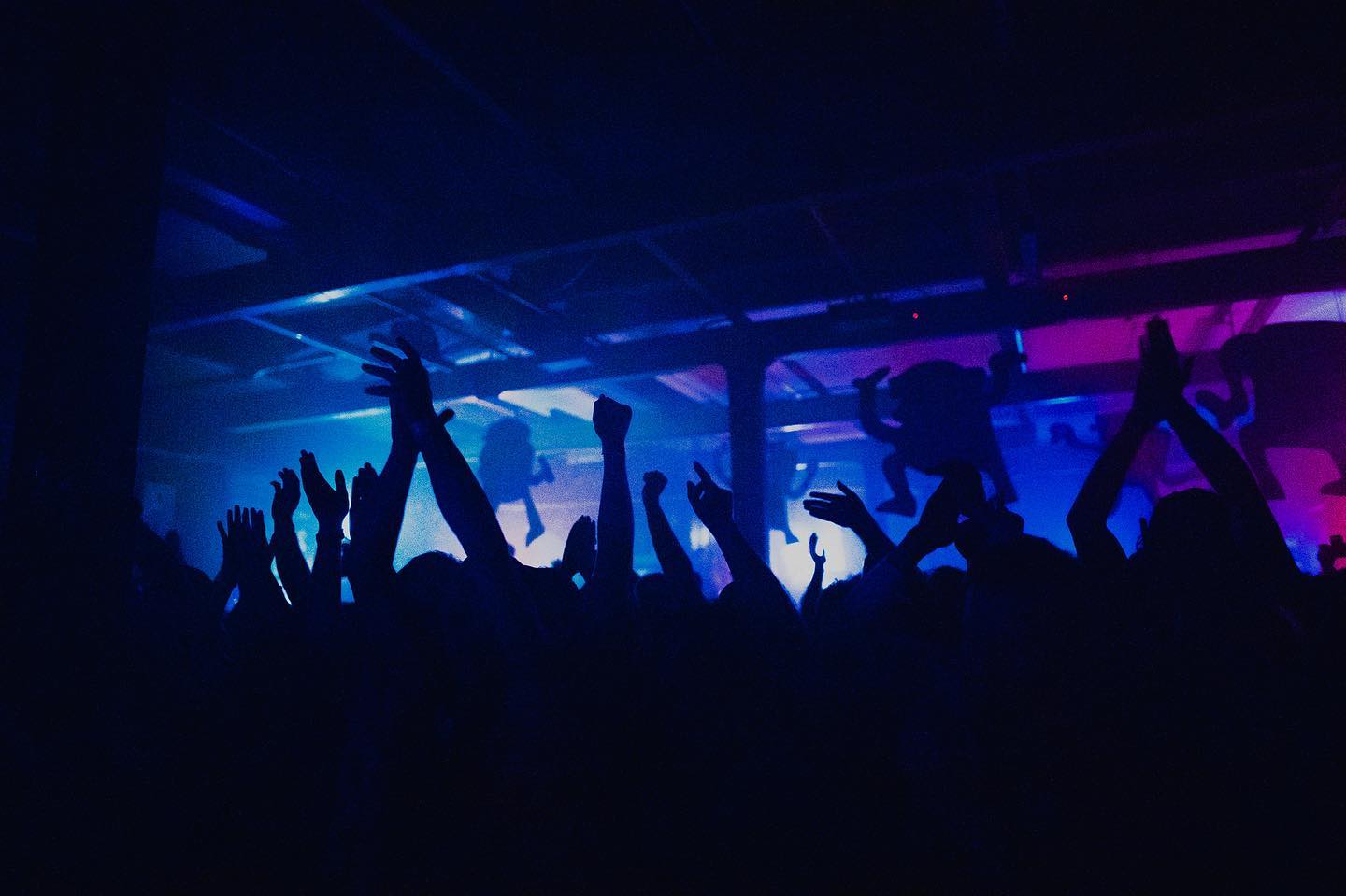 A crowd dances, arms held high, at a night club. The lighting is all blues and purples.