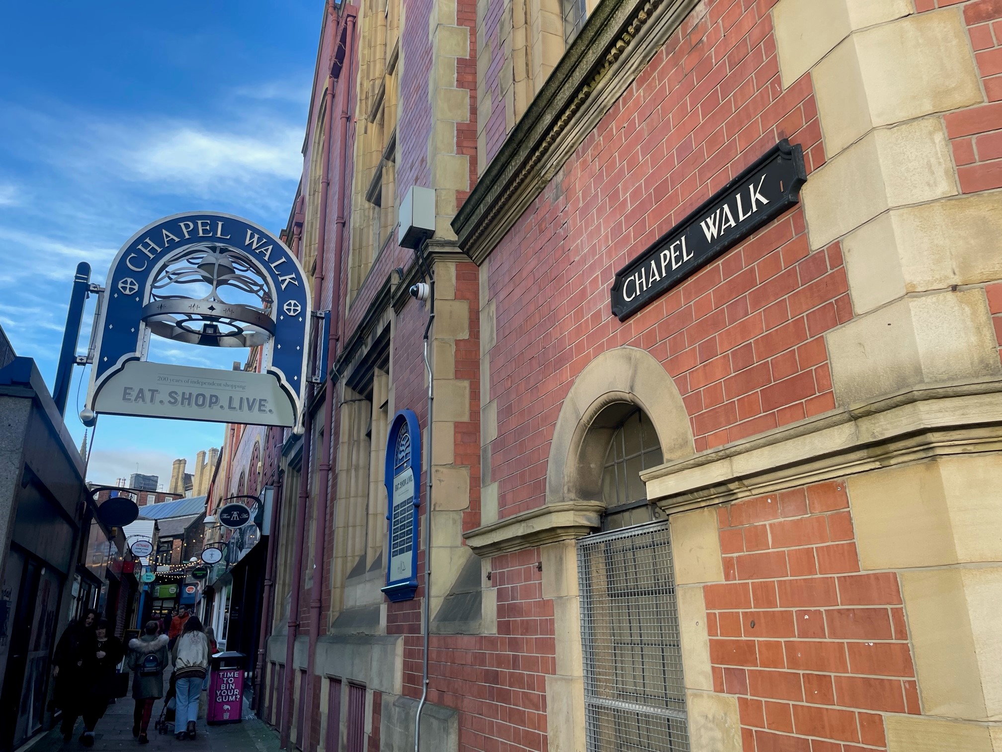 Narrow pedestrian walkway called Chapel Walk, lined with red brick buildings featuring stone detailing. A black sign reading ‘Chapel Walk’ is mounted on the wall, and a decorative hanging sign with the same name and the words ‘Eat. Shop. Live.’ is visible above the entrance. Several smaller shop signs and people walking can be seen further down the alley under a bright blue sky.