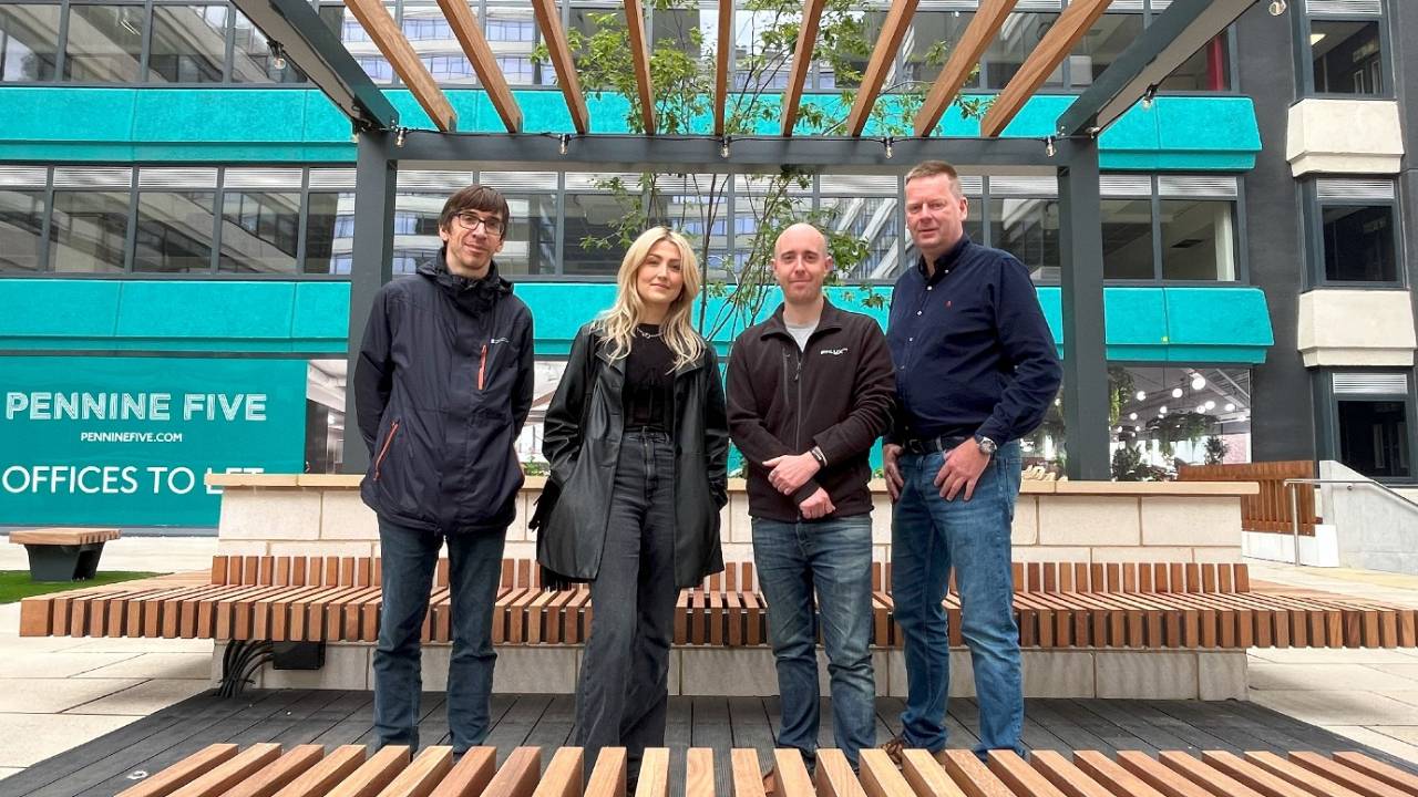 Four people standing together in an outdoor courtyard area with modern architecture. The setting features wooden benches arranged in rows, a pergola-style structure overhead, and a small tree planted in the center. Behind them is a multi-story building with teal panels and large windows, displaying signage that reads “Pennine Five” and “Offices To Let.”