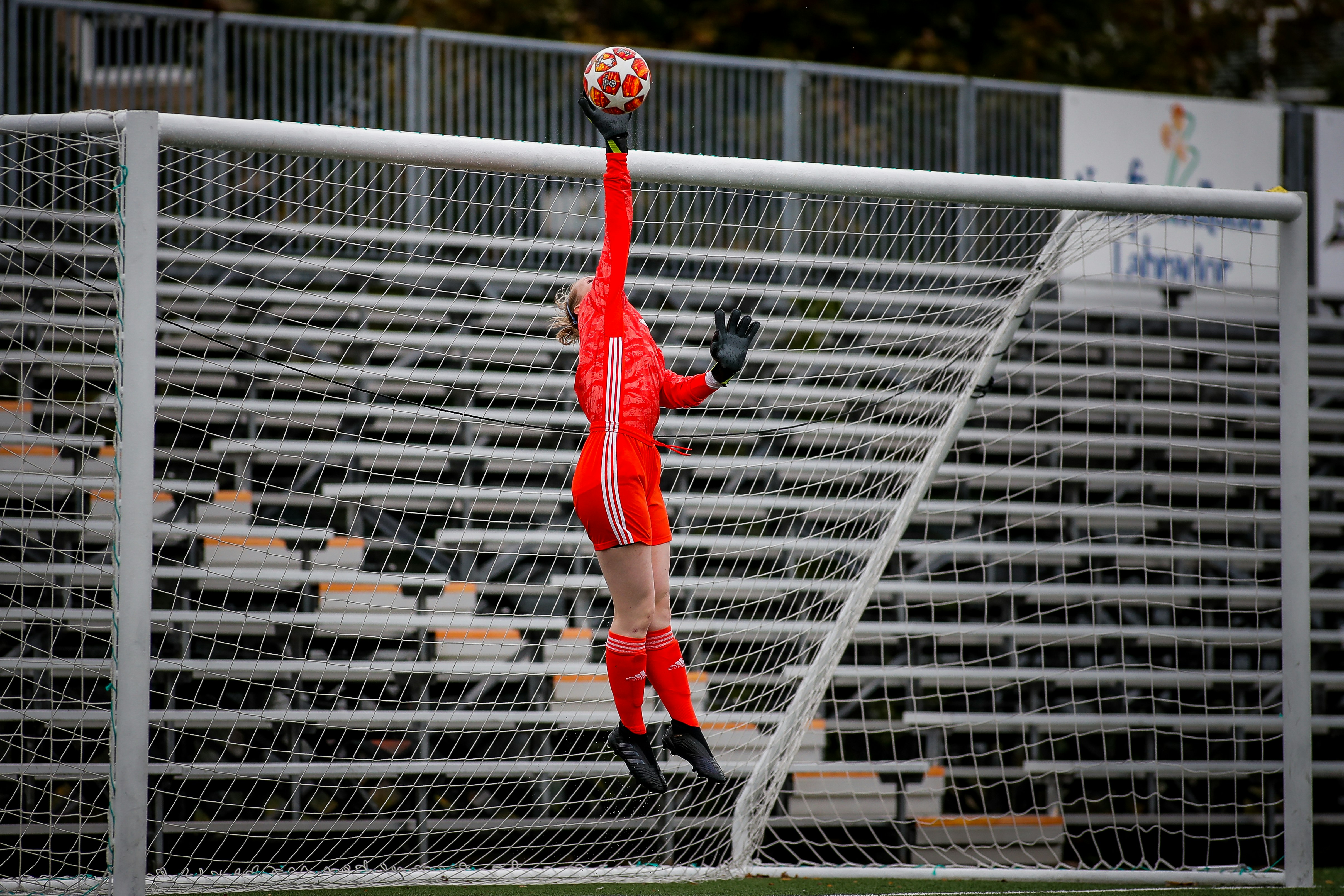 Panoramic view of a packed football stadium with fans filling the stands. The green pitch is set for a match, with goalposts visible at both ends. Large red banners and flags are displayed by supporters, and the stadium roof arches overhead with a modern design.