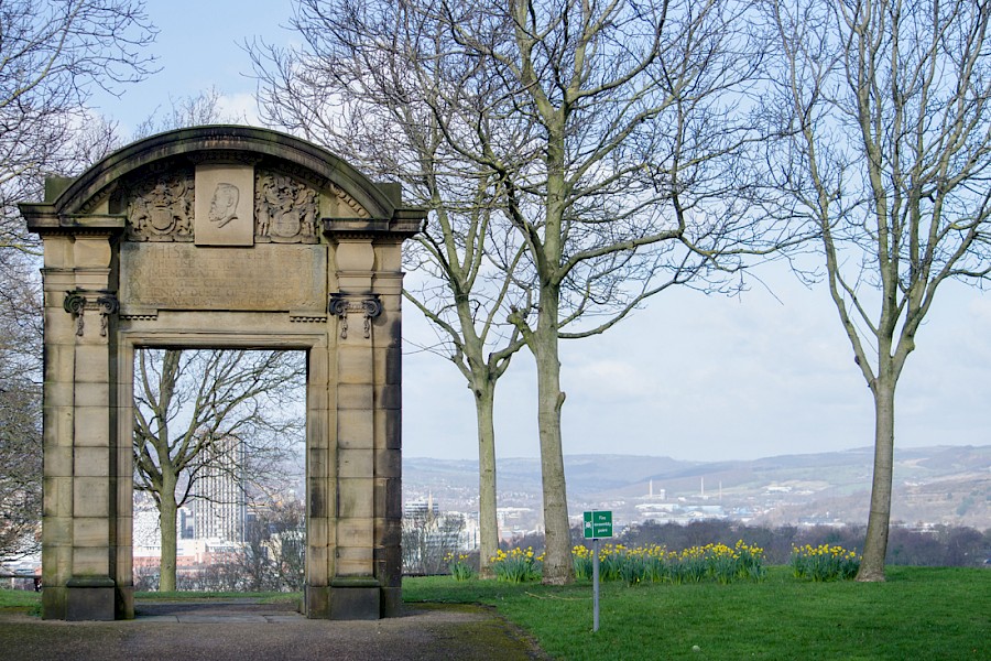 A historic stone archway with decorative carvings stands in a park surrounded by bare trees. The arch frames a distant view of a cityscape with hills in the background. Yellow daffodils bloom in the grassy area near the arch, and a small green sign is visible on the lawn. The sky is bright with scattered clouds, creating a clear and open atmosphere.