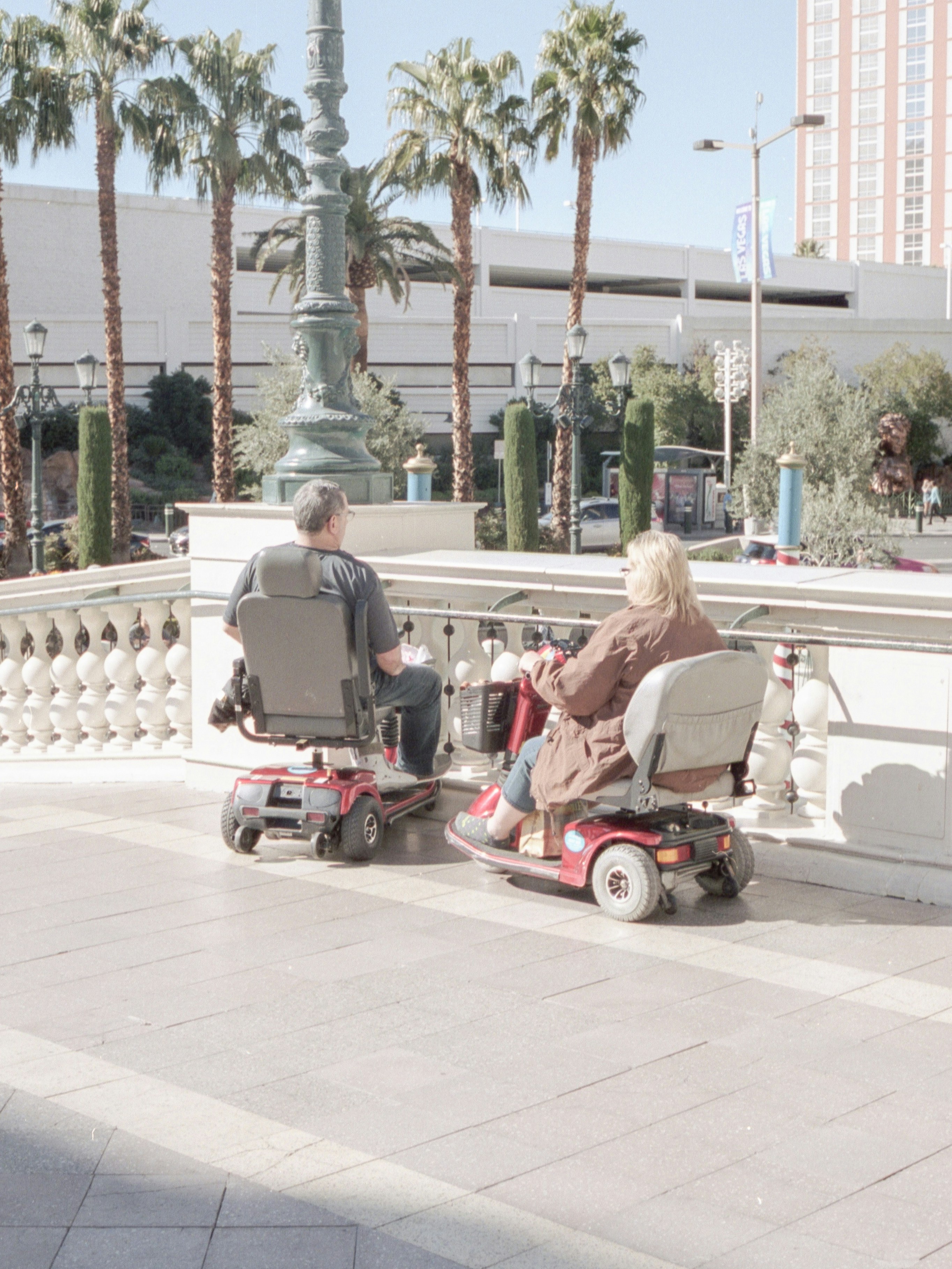 Two people seated on mobility scooters on a paved outdoor area, facing a decorative stone railing. Behind them are tall palm trees, a large ornate street lamp, and a multi-story building in the background under bright daylight