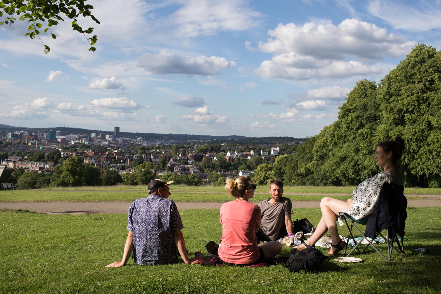 A group of four people, sat in Meersbrook Park, enjoying a sunny day. One is sat in a folding chair and the other three are sat on the grass.
