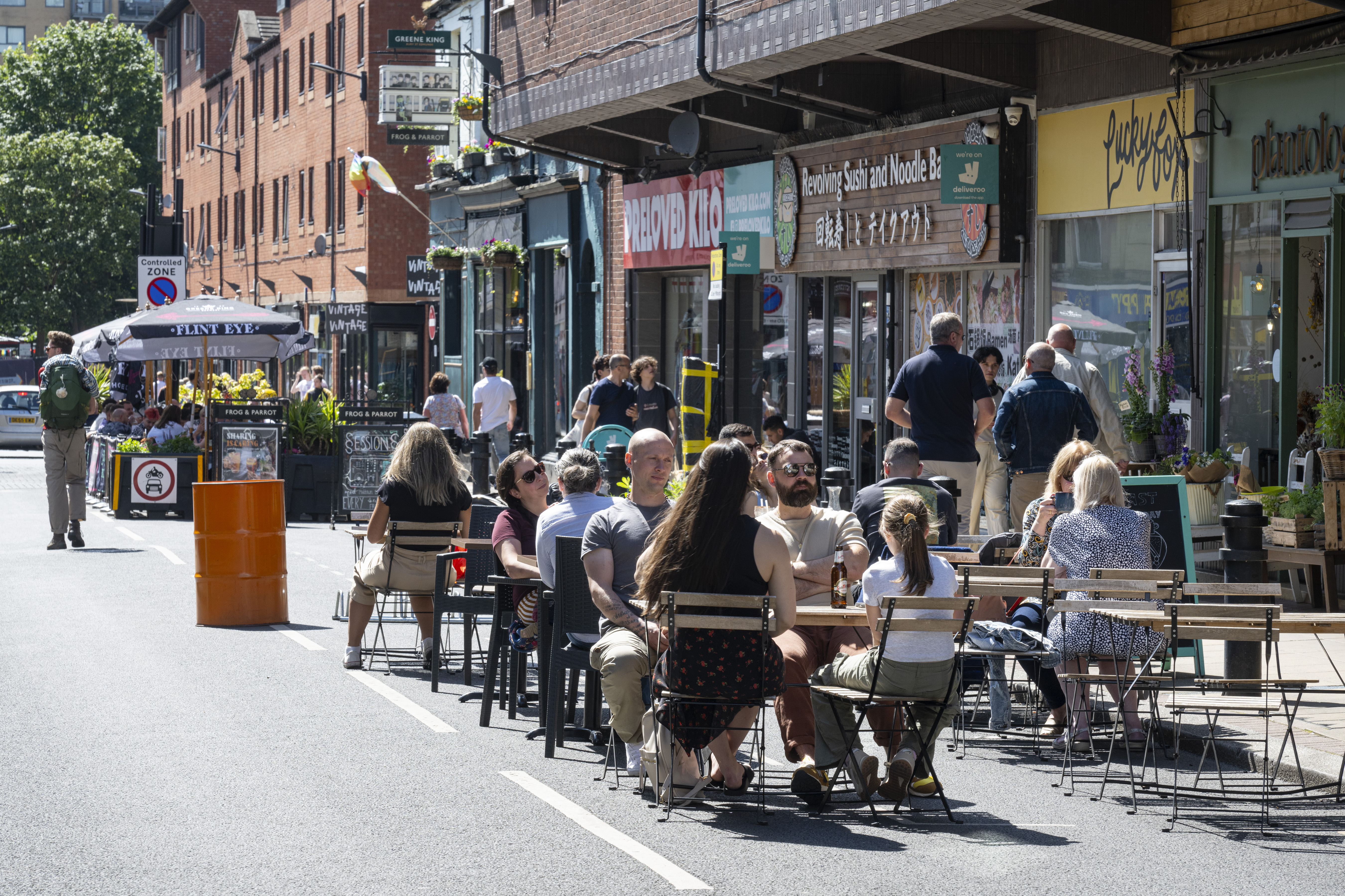 People are seated at outdoor tables on a closed-off city street, enjoying food and drinks in sunny weather. The street is lined with shops and restaurants, including signs for a noodle bar and a café. A bright orange barrel and potted plants decorate the area, while pedestrians walk along the street in the background. The scene conveys a lively, social atmosphere in an urban setting.