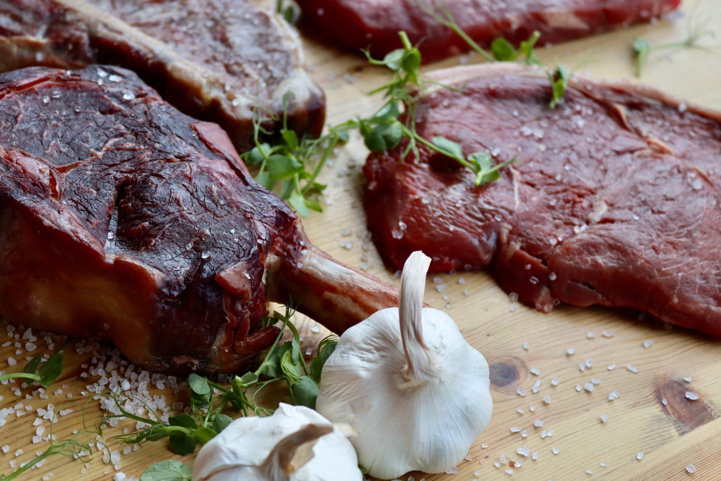 Several cuts of meat on a chopping board with bulbs of garlic and herbs.