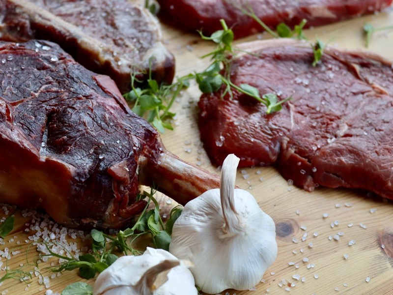 Several cuts of meat on a chopping board with bulbs of garlic and herbs.