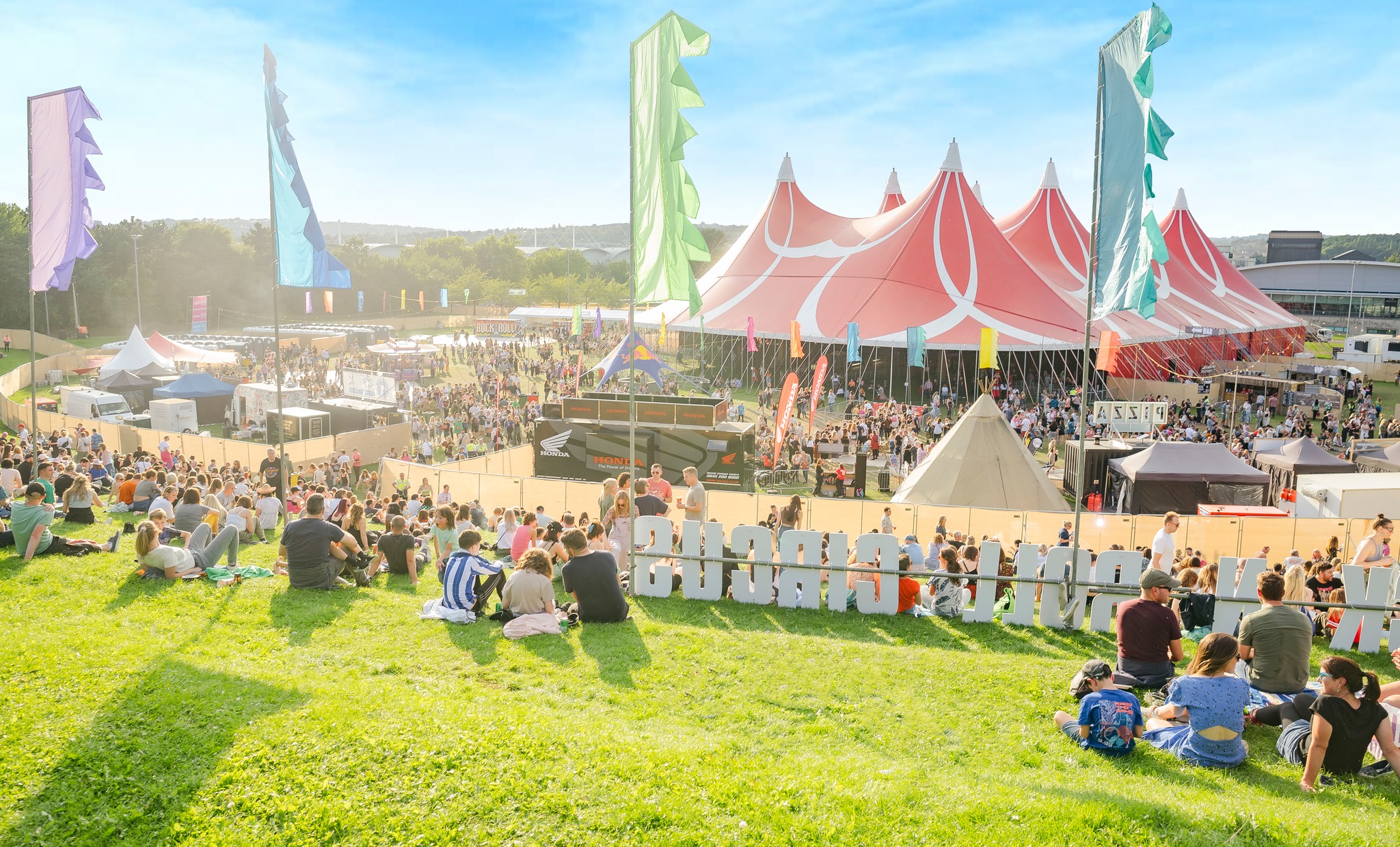 Large outdoor music festival with crowds gathered on a grassy hill and around a big red-and-white striped tent. Colorful vertical flags line the area, and smaller tents and stalls are visible in the background under a bright blue sky.
