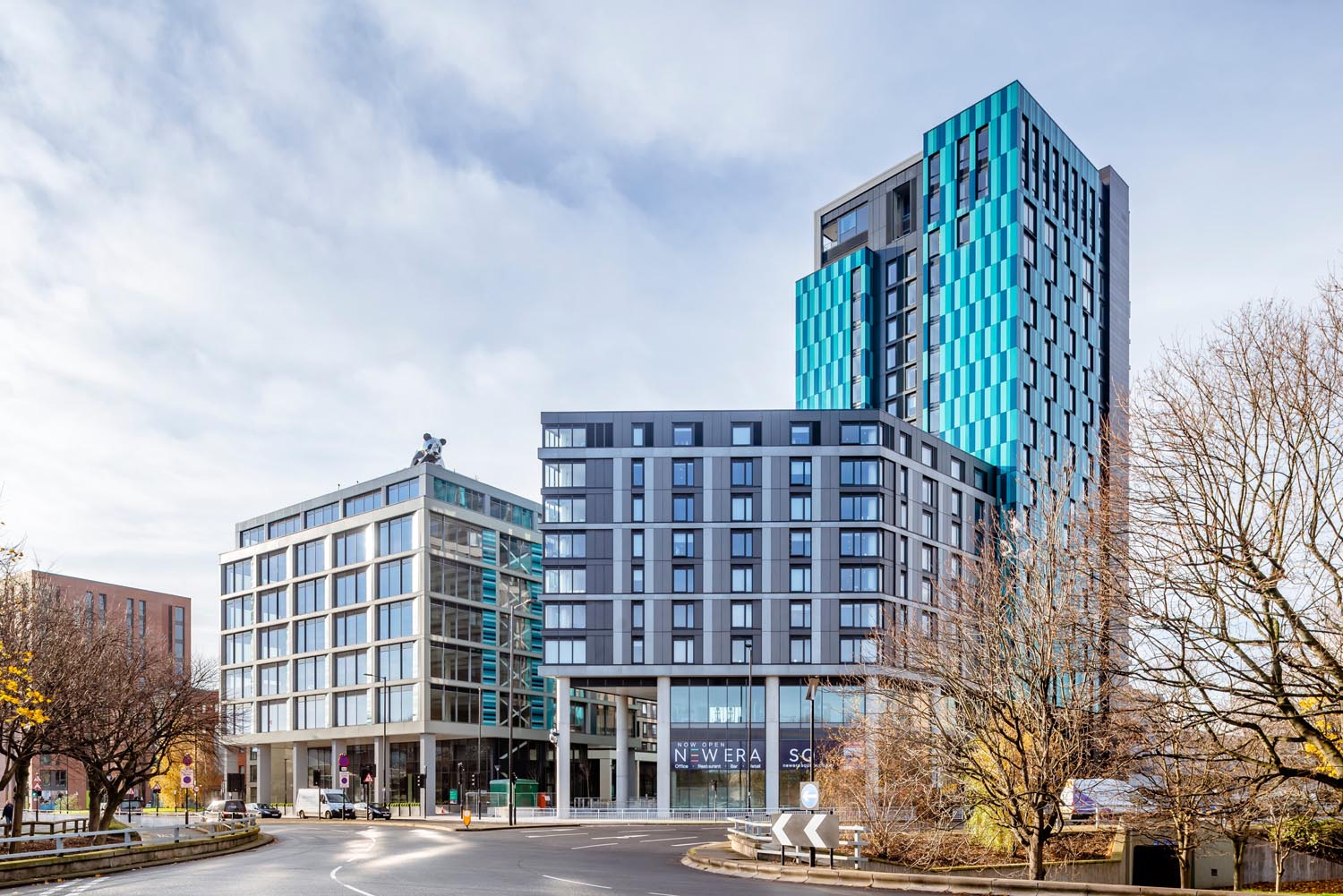Modern cityscape featuring two contemporary high-rise buildings at a roundabout. The left building has a glass façade with a grid-like design, while the taller building on the right has a striking blue and gray exterior with geometric patterns. Bare trees frame the scene, and signage at street level reads “New Era Square.” The sky is partly cloudy, creating a bright and open atmosphere.