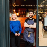 Two people standing in the doorway of a cozy restaurant or café. One is wearing a bright blue long-sleeve top and dark pants, while the other is dressed in a black shirt with a dark apron tied at the waist. The interior behind them features warm lighting, shelves with books, and string lights. A handwritten orange sign is partially visible inside, and the exterior has dark framing around the entrance.