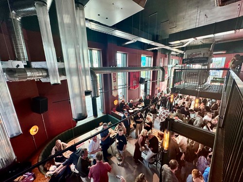 Interior view of a busy bar or restaurant with an industrial-style design, featuring exposed metal pipes, high ceilings, and large windows letting in natural light. The space is filled with people standing and sitting at tables, socializing and enjoying drinks. Warm lighting from wall lamps and hanging fixtures adds to the lively atmosphere.