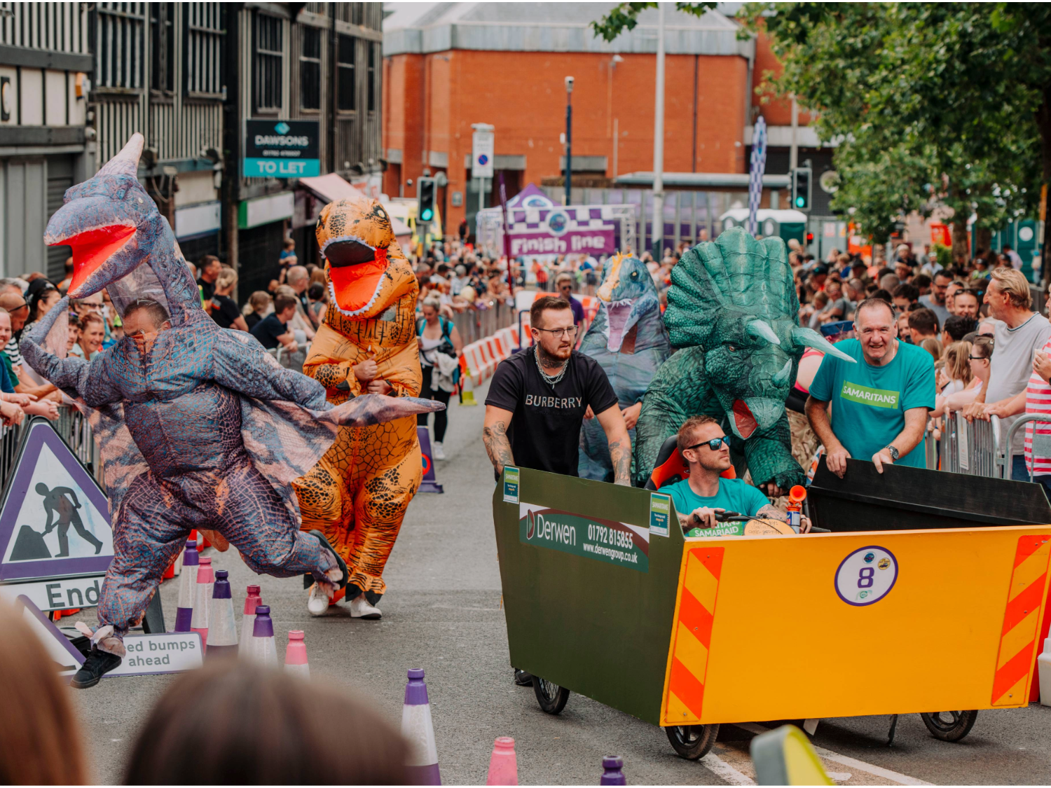 A group of participants in colorful dinosaur costumes run alongside a large yellow soapbox cart with orange chevron markings during a city street race. The cart has event branding and is surrounded by purple-and-white cones and red-and-white barriers. Crowds of spectators line both sides of the track, and a purple “Finish line” banner is visible in the background along with buildings and event signage.