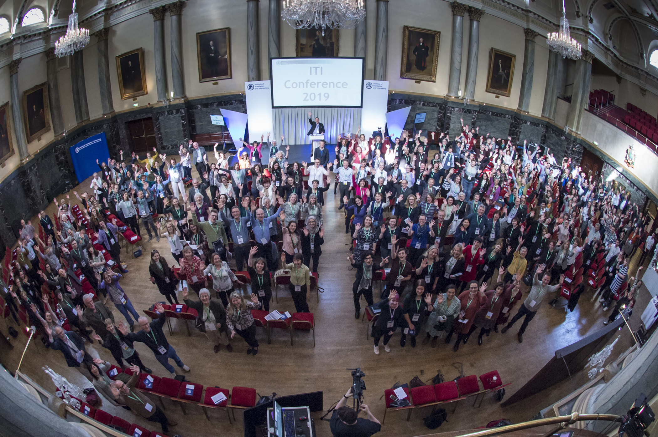 A large group of people gathered in a grand hall for the ITI Conference 2019. The hall features ornate architectural details, chandeliers, tall columns, and framed portraits on the walls. Attendees are standing and facing the stage, where a speaker is positioned at a podium beneath a screen displaying the conference name. Red chairs are arranged in rows, and a camera setup is visible in the foreground capturing the event.