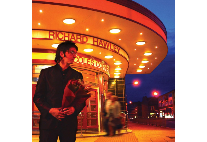 A person dressed in a dark suit stands outside an illuminated art deco-style building at night, holding a bouquet of flowers. The building features a curved canopy with round lights and signage reading “Richard Hawley” and “Coles Corner.” The background shows a deep blue evening sky and blurred figures walking near the entrance.