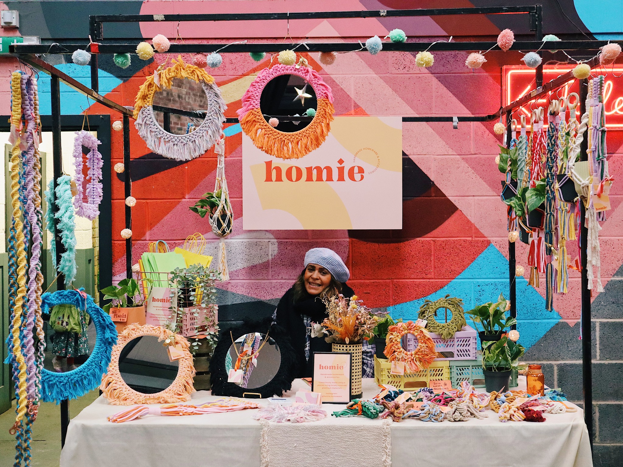A woman is sat smiling behind a stall, selling knitted and crocheted goods.