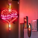 A cheeky neon sign that shows a heart pierced by a dagger with the words 'Sex, drugs & bacon rolls' on the wall at The Riverside Kelham.