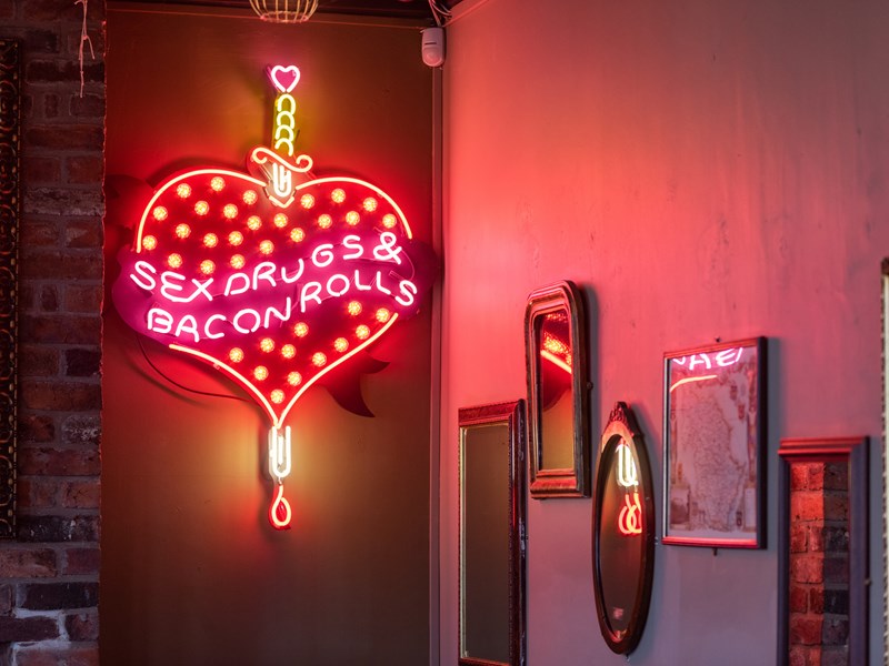 A cheeky neon sign that shows a heart pierced by a dagger with the words 'Sex, drugs & bacon rolls' on the wall at The Riverside Kelham.