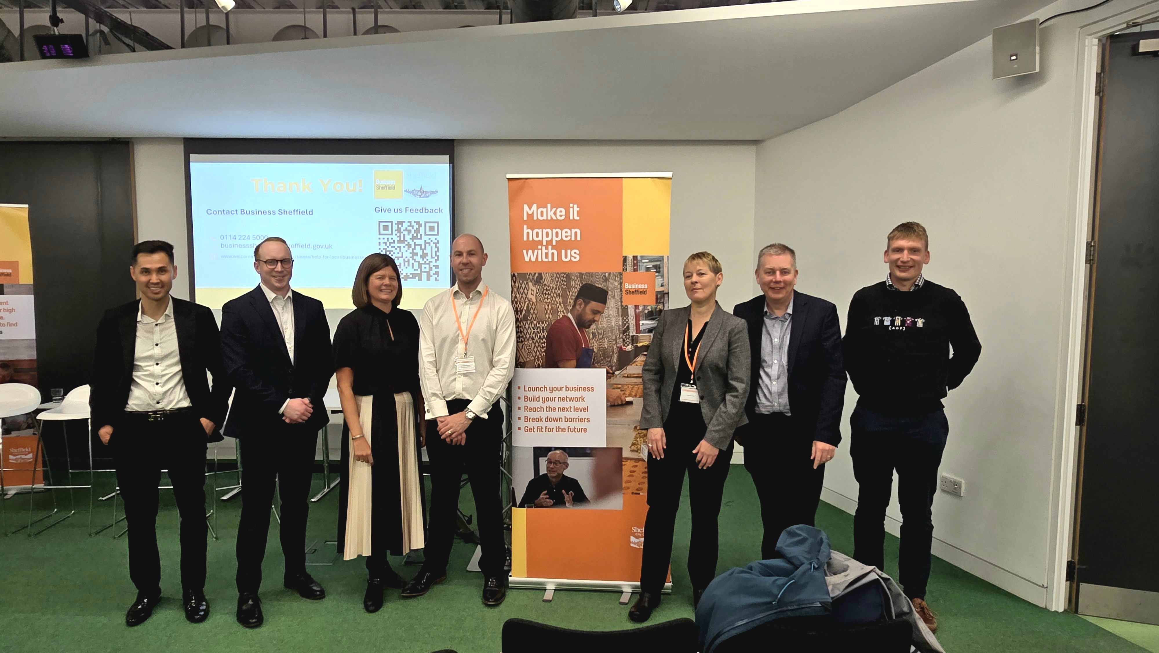 Group of seven people standing indoors in front of a presentation screen and a tall orange banner. The banner reads ‘Make it happen with us’ and includes text about business support along with an image of a person working on a laptop. The presentation screen behind them displays a thank-you message with contact details and a QR code. The setting appears to be a professional event or networking session with green carpet flooring and white walls.
