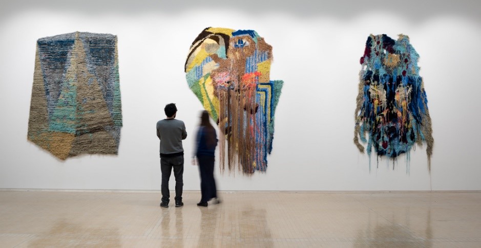 Two people standing in a gallery observing three large textile artworks mounted on a white wall. The pieces feature abstract designs with layered textures and vibrant colors, including shades of blue, green, yellow, and brown. The central artwork has long strands hanging down, creating a dynamic effect. The floor is light-colored and reflective, and the overall setting is minimalistic, emphasizing the art.