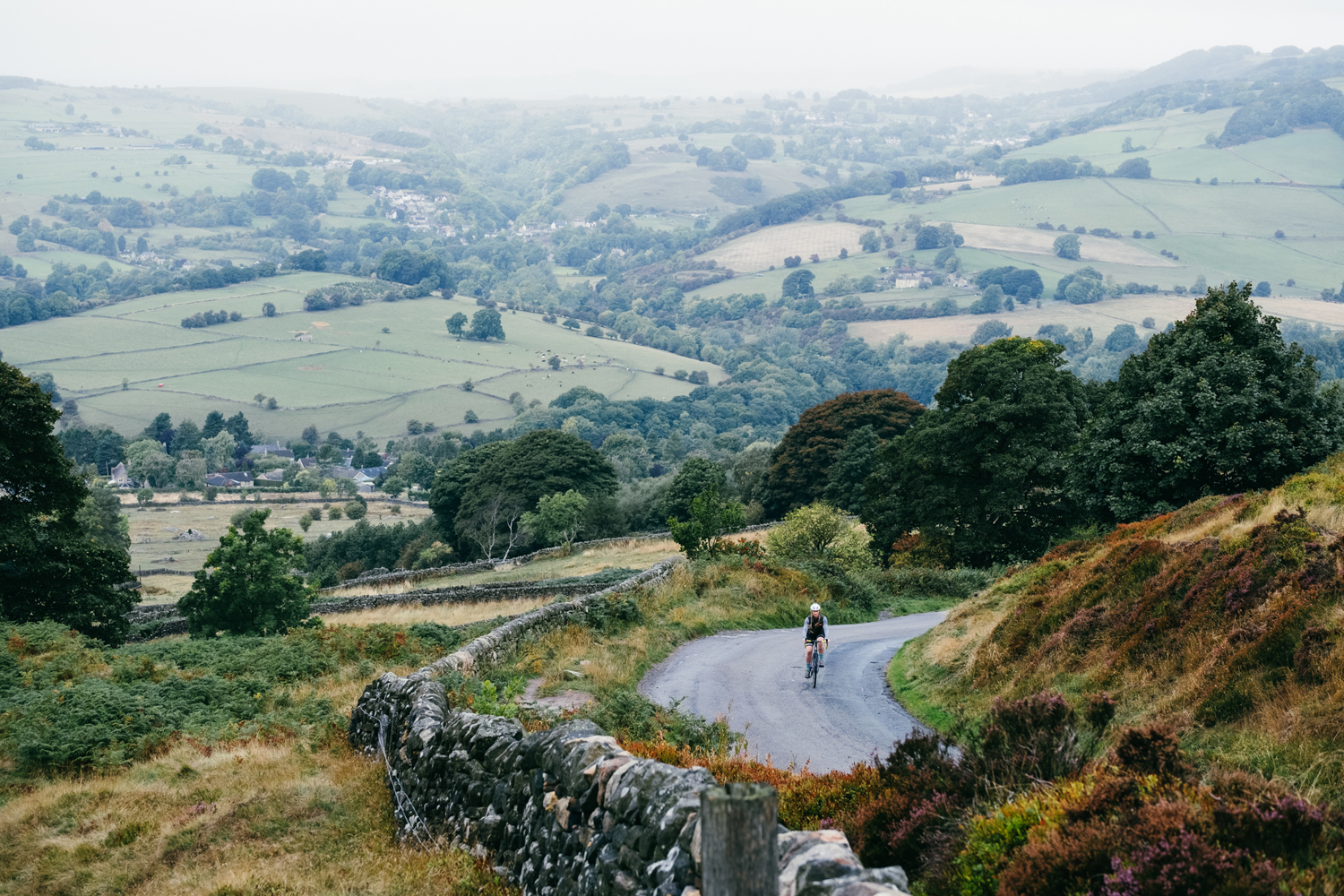 Aerial shot of cyclist riding along countryside road with a stone wall, further fields of countryside in the background.