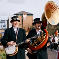 Two musicians, one playing a banjo and the other a brass sousaphone, perform at an outdoor market in Sheffield.