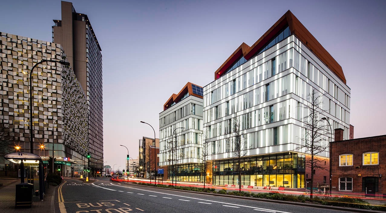 Modern city street at dusk with contemporary glass buildings featuring angular rooftops and illuminated interiors. On the left, a tall patterned building contrasts with the sleek design of the two main structures. The road in the foreground has bus lane markings, streetlights, and light trails from passing vehicles, with a clear sky in the background.