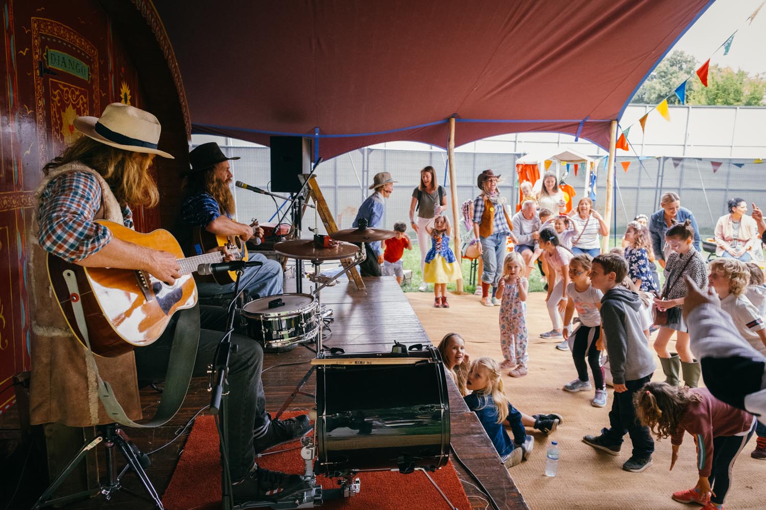 A lively scene under a large tent where two musicians are performing on a small stage. One musician is playing an acoustic guitar while the other is seated at a drum set. In front of the stage, a group of children and adults are gathered, some dancing and others watching the performance. The area is decorated with colourful bunting, and the background shows outdoor structures and greenery, suggesting a festival or community event.