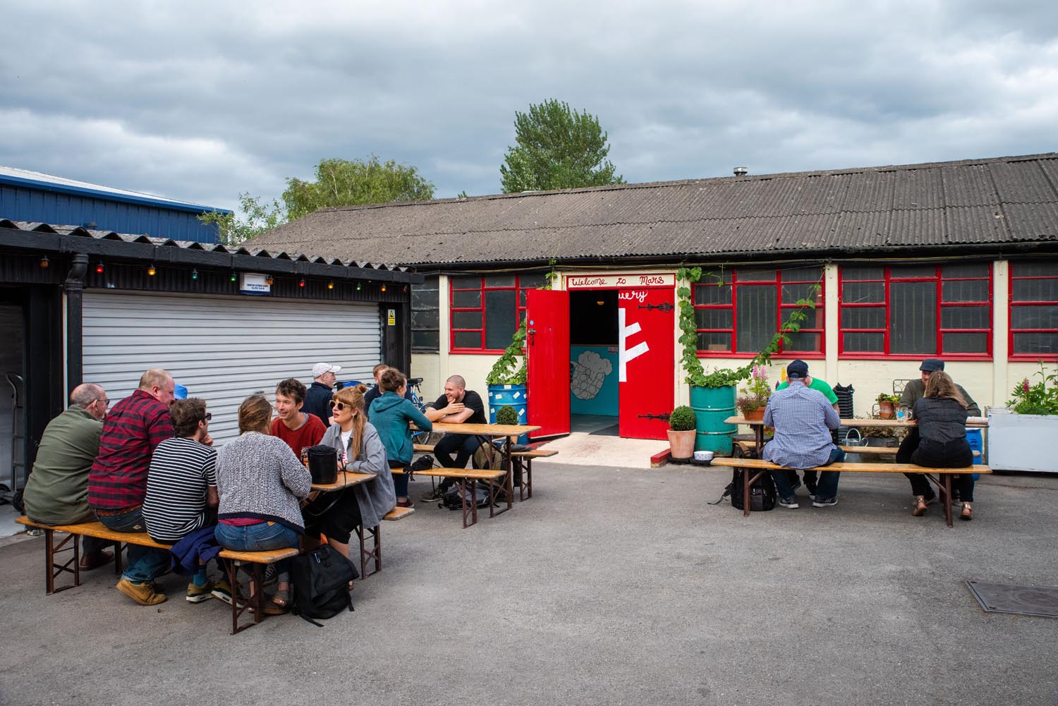 Outdoor seating area with wooden picnic tables and benches, occupied by groups of people. A rustic building with red-framed windows and a red door is in the background, with potted plants near the entrance under an overcast sky.