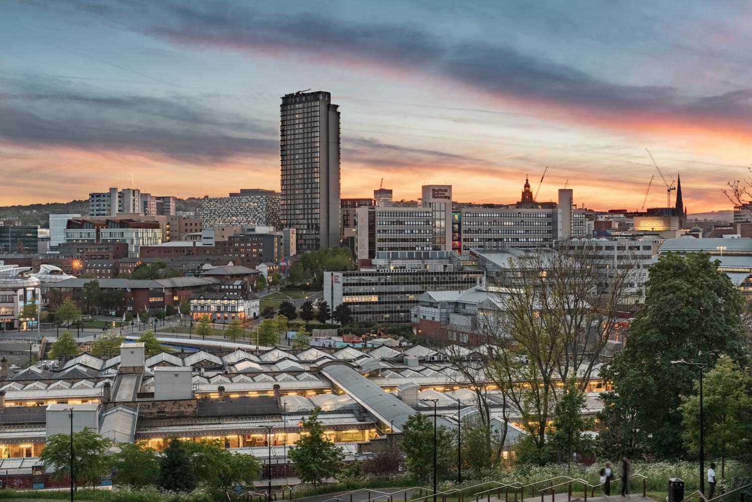A panoramic view of Sheffield city at sunset, featuring a mix of modern and historic architecture. In the foreground, rooftops of a large building complex with angled glass panels are visible. The middle ground shows office buildings and residential blocks, while the background includes the prominent tall tower of the city center and several cranes. The sky is painted with warm orange and pink hues blending into blue, creating a striking backdrop for the urban landscape.