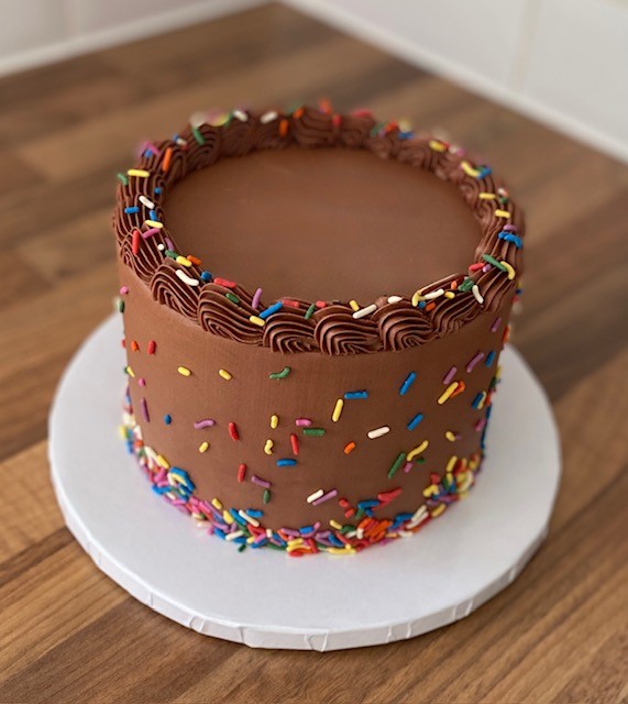 A round vegan chocolate cake covered in smooth chocolate frosting, decorated with colorful rainbow sprinkles around the sides and top edge. The cake sits on a white cake board on a wooden surface.