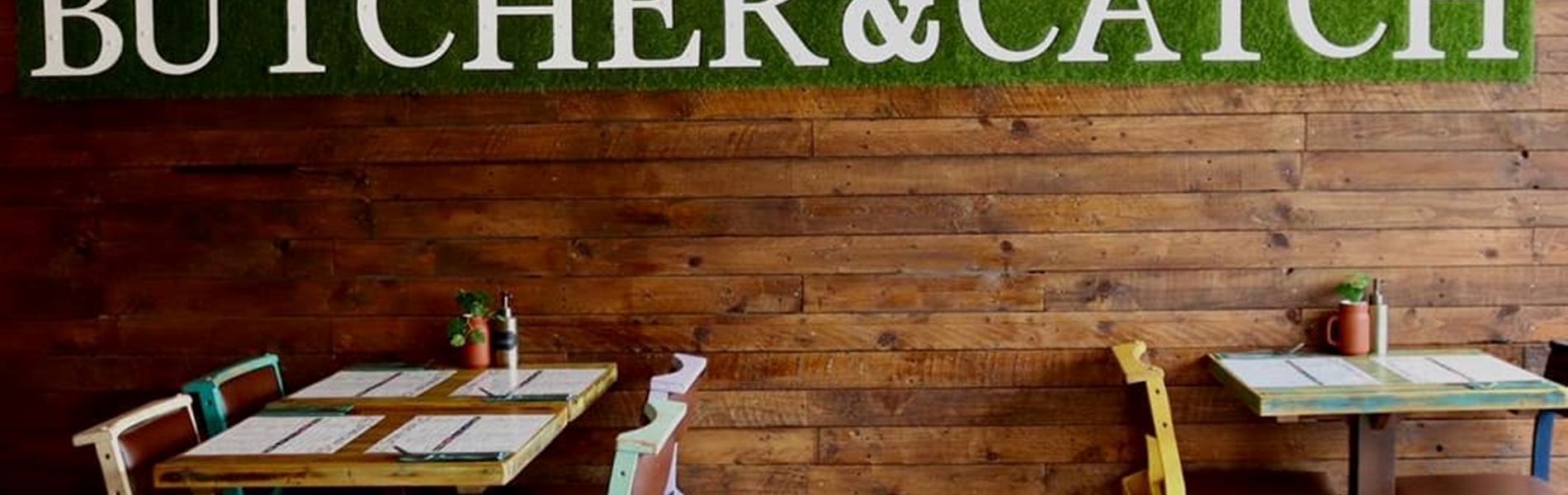 Restaurant tables next to a naked brick wall with a green sign that reads 'Butcher & Catch'.