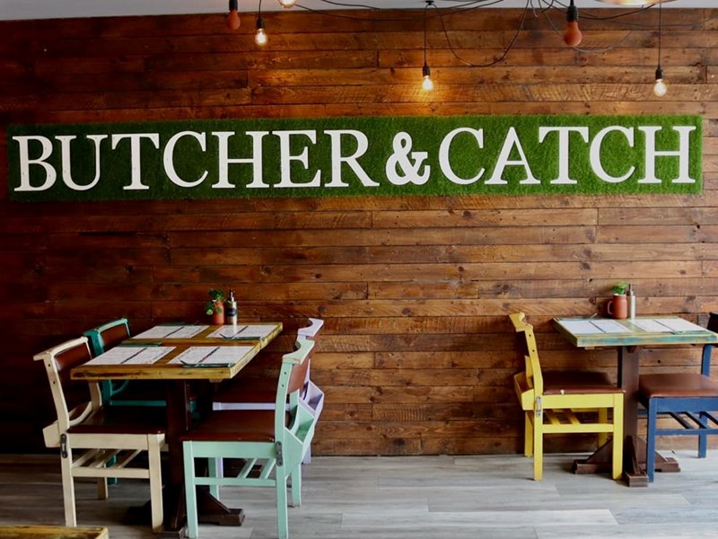 Restaurant tables next to a naked brick wall with a green sign that reads 'Butcher & Catch'.