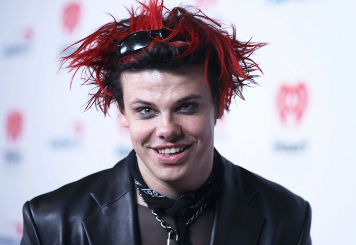 Person wearing a black leather jacket and layered necklaces, including a padlock pendant, with a black scarf tied around the neck. Hair is styled in spiky red and black strands with sunglasses resting on top. The background features a white backdrop with red heart-shaped logos.