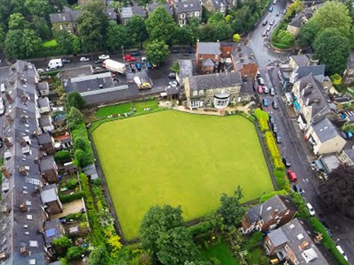 Aerial view of a rectangular green lawn, possibly a bowling green, surrounded by residential houses and narrow streets. The area includes trees, gardens, and parked cars, with a mix of brick buildings and greenery visible throughout the neighbourhood.