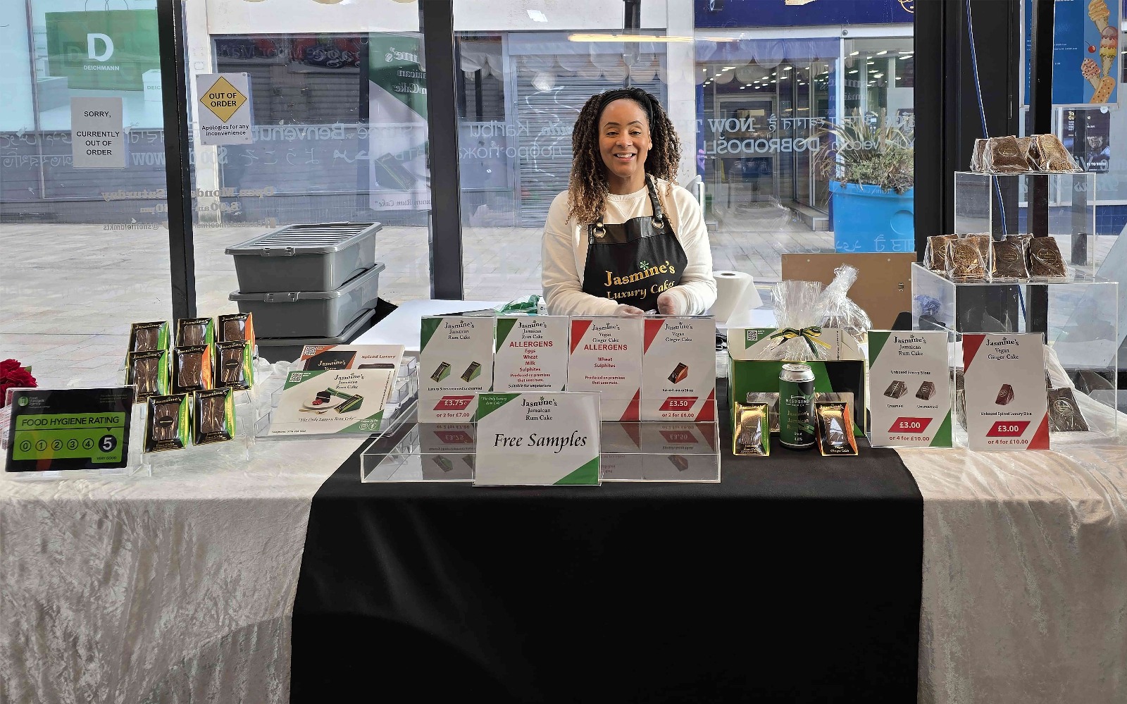 A display table set up for a food sampling event inside a bright indoor space with large glass windows. The table is covered with a black and white cloth and features neatly arranged packaged snacks, promotional signs, and a tray labeled “Free Samples.” There are clear acrylic stands holding additional products on the right side, and a small green sign with food hygiene rating on the left.