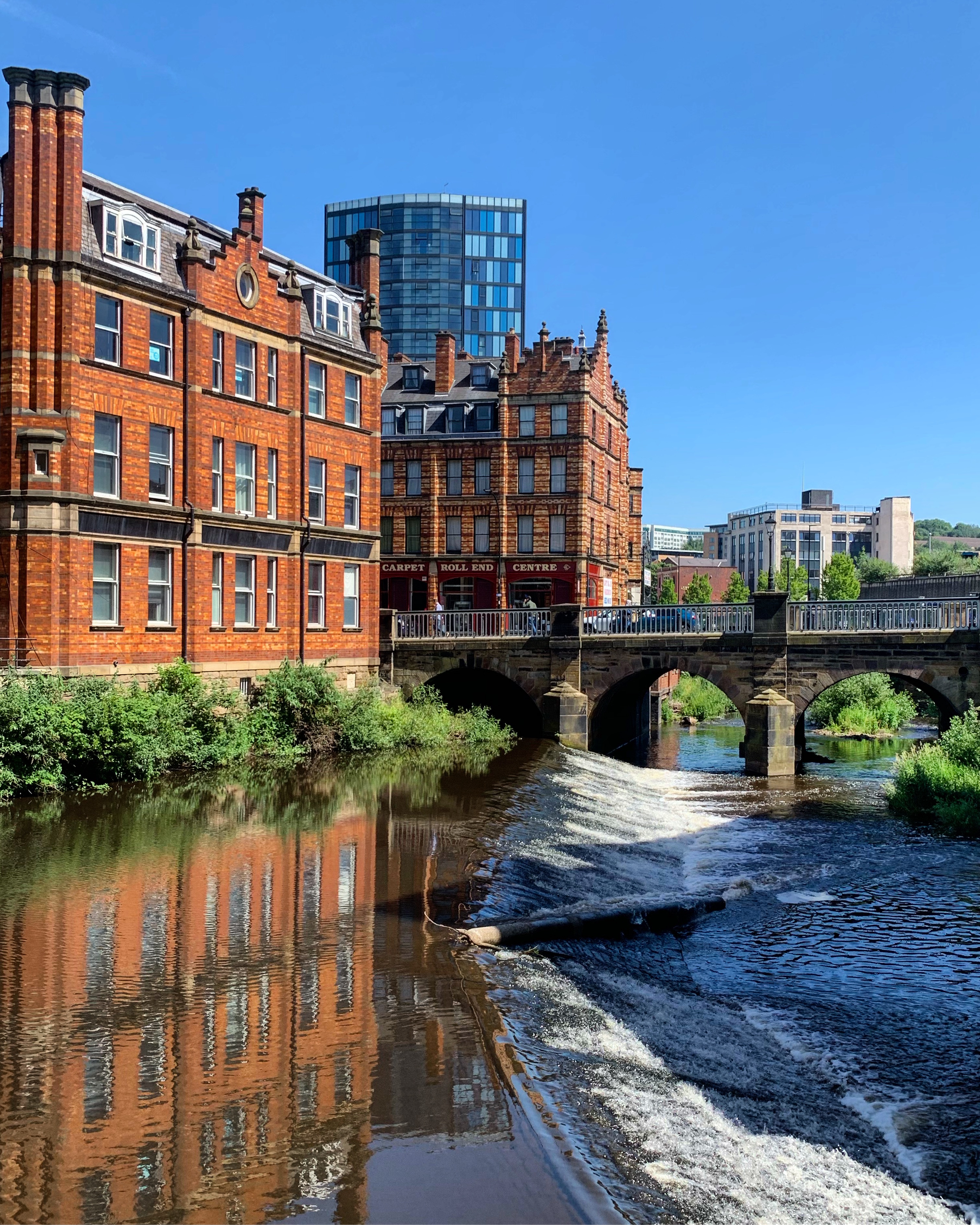 A scenic view of a river flowing through a city with a small weir in the foreground. On the left, there are historic red-brick buildings with ornate architectural details, reflected in the calm water. A stone bridge with three arches spans the river, and behind it rises a modern glass high-rise building against a clear blue sky. Green shrubs line the riverbank, adding a touch of nature to the urban setting.