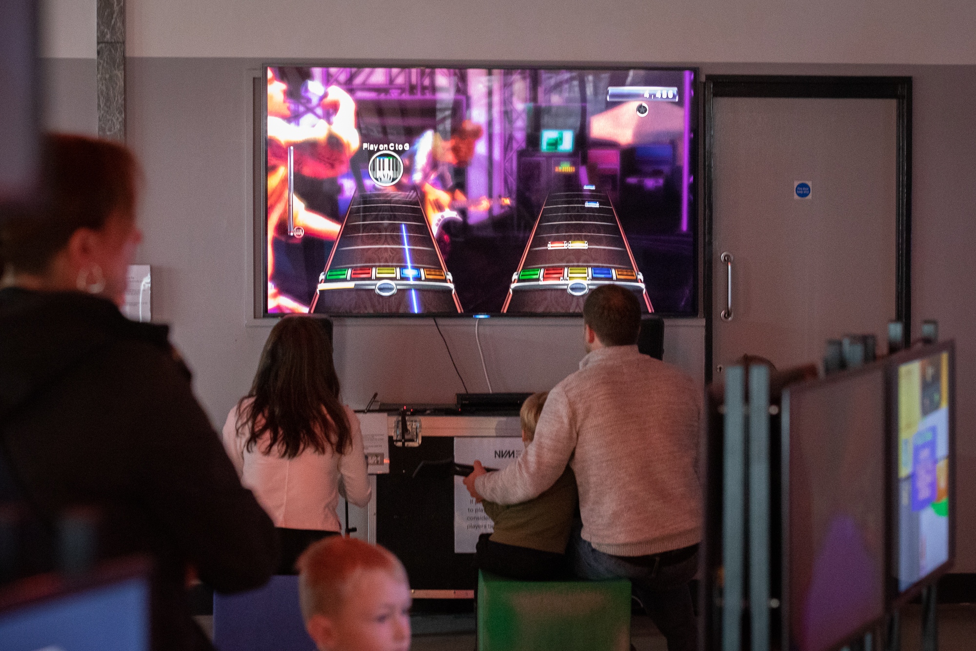 Two children and an adult are playing a video game at the National Videogame Museum. Other people are looking at exhibitions.