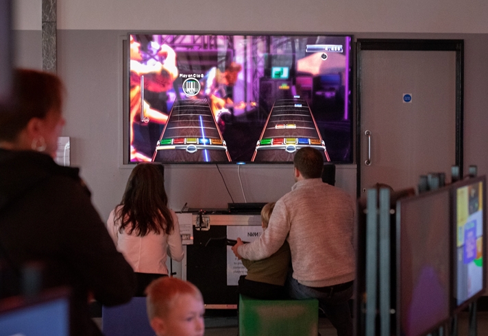 Two children and an adult are playing a video game at the National Videogame Museum. Other people are looking at exhibitions.