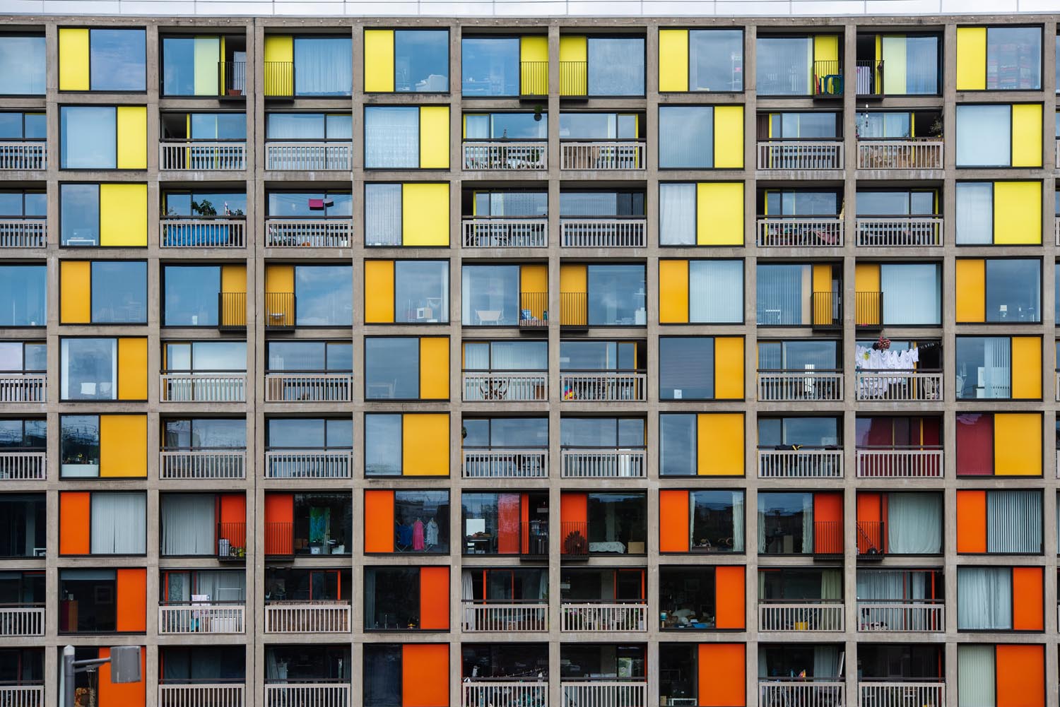 Front view of a multi-story residential building with a grid-like facade featuring large windows and panels in shades of yellow, orange, and red. Each unit has a balcony with railings, and some balconies display hanging laundry or personal items.