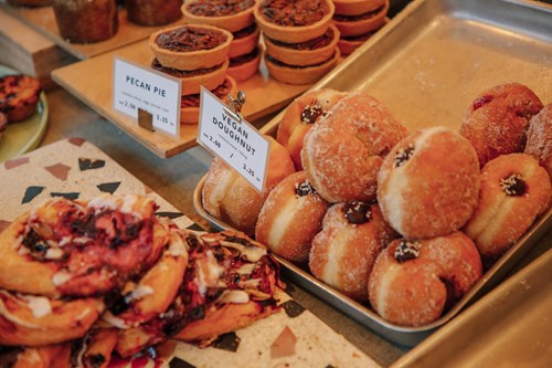 Display of assorted baked goods on a counter, including a tray of sugar-coated vegan doughnuts filled with jam, pecan pies arranged on a wooden board, and fruit pastries with a glossy finish. Small signs indicate ‘Pecan Pie’ and ‘Vegan Doughnut’ with prices.