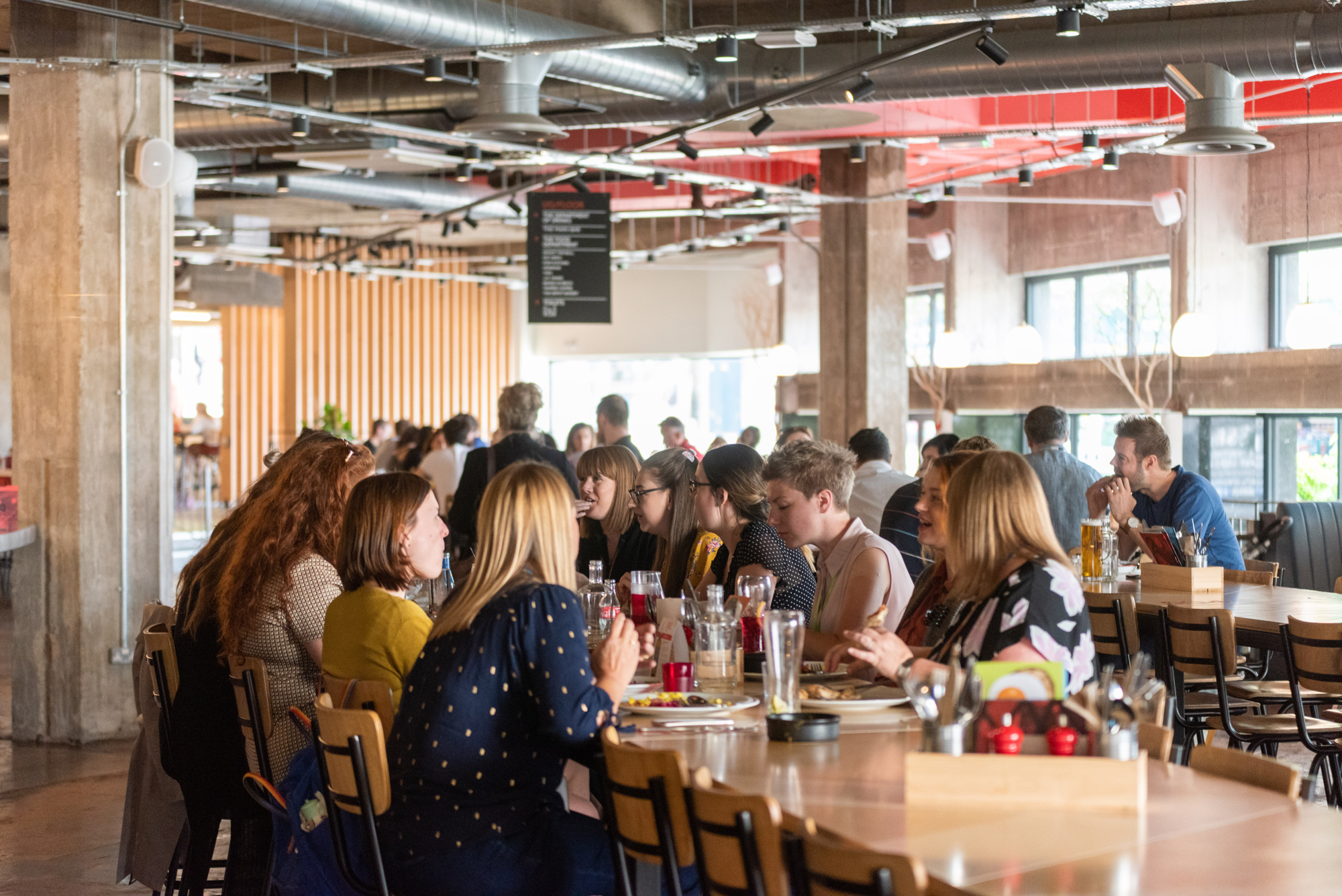 Kommune Food Hall - Interior of a modern food hall with industrial-style design, featuring exposed ducts, concrete pillars, and large windows. Several groups of people are seated at long wooden tables enjoying food and drinks. The space is well-lit with natural light and hanging ceiling lights, and a black menu board is visible in the background.