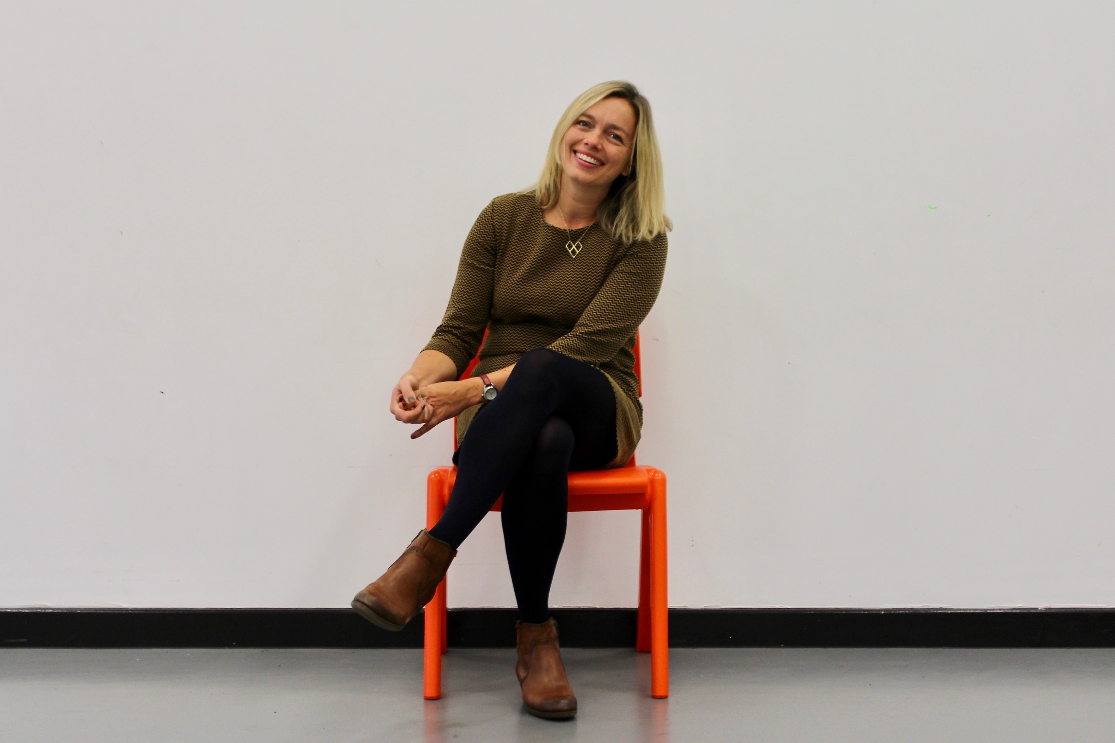 A person seated on a bright orange chair against a plain white wall. The individual is wearing a textured brown long-sleeve dress, dark leggings, and brown ankle boots. Hands are loosely clasped, and legs are crossed at the ankles. The setting appears to be indoors with a smooth gray floor and minimal background detail.