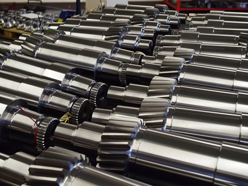 Close-up view of multiple shiny metal gears and cylindrical components arranged in rows. The parts have polished surfaces with visible teeth and grooves, suggesting precision engineering for industrial machinery or mechanical systems.